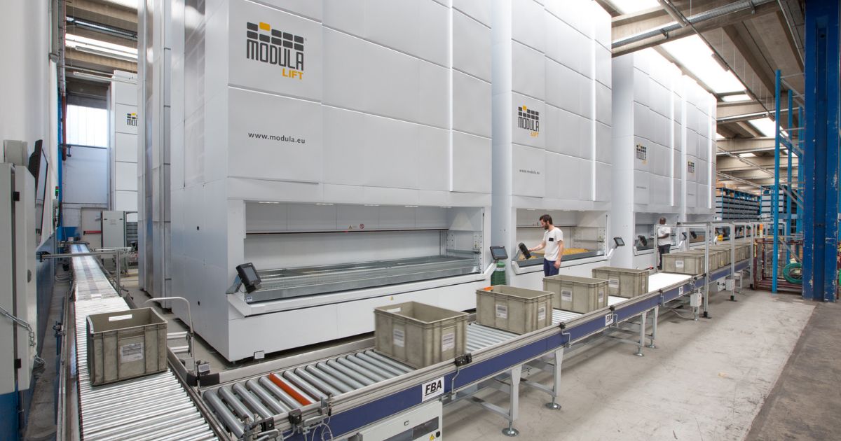 A worker operates a Modula lift machine with a conveyor belt in a warehouse setting.